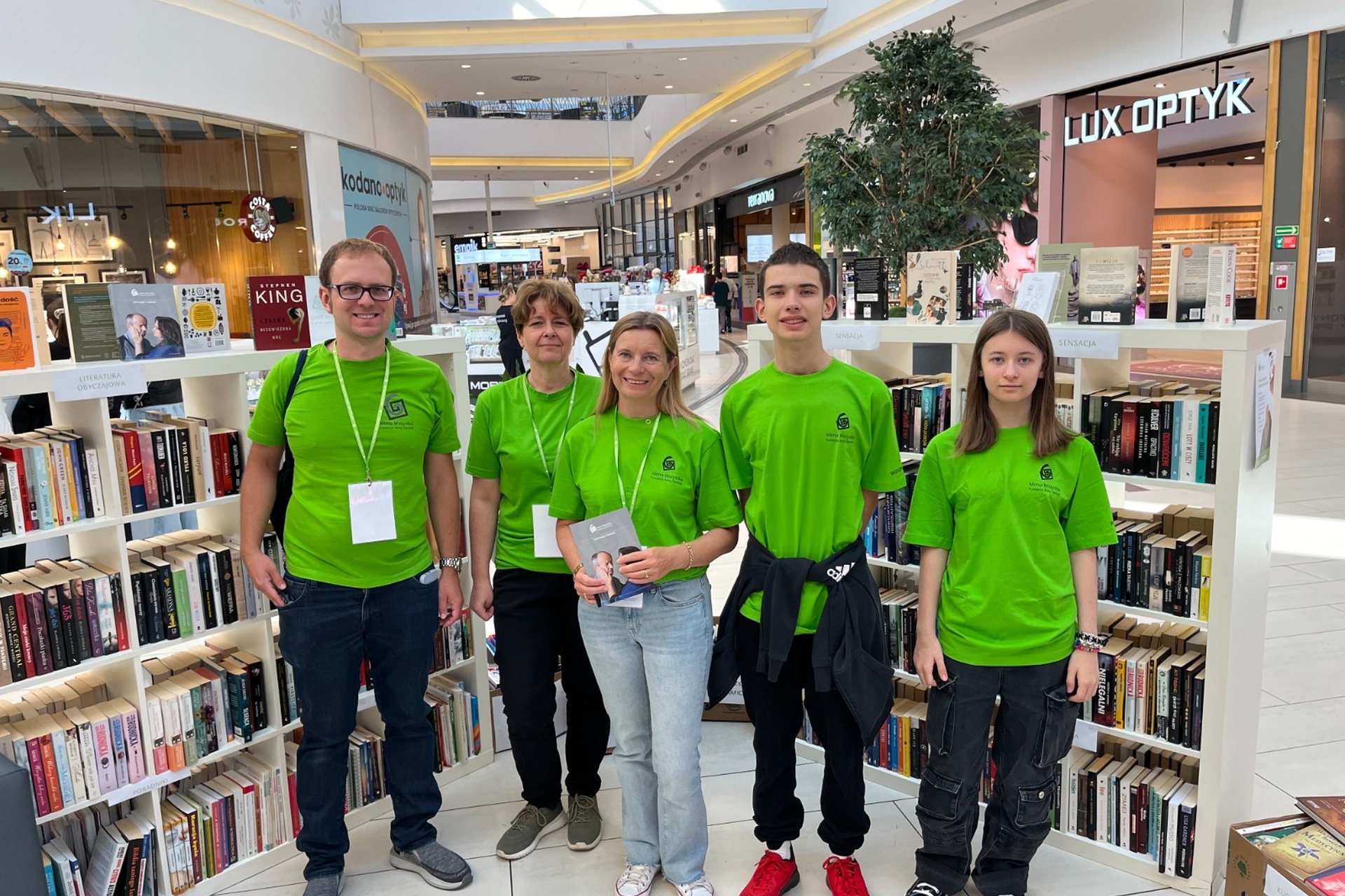 5 osób w różnym wieku stoi na środku, zwrócona twarzami. Są ubrani w zielone koszulki i długie spodnie. Wszyscy się uśmiechają. Jedna kobieta trzyma w ręce książkę. Za nimi widać regały wypełnione książkami.