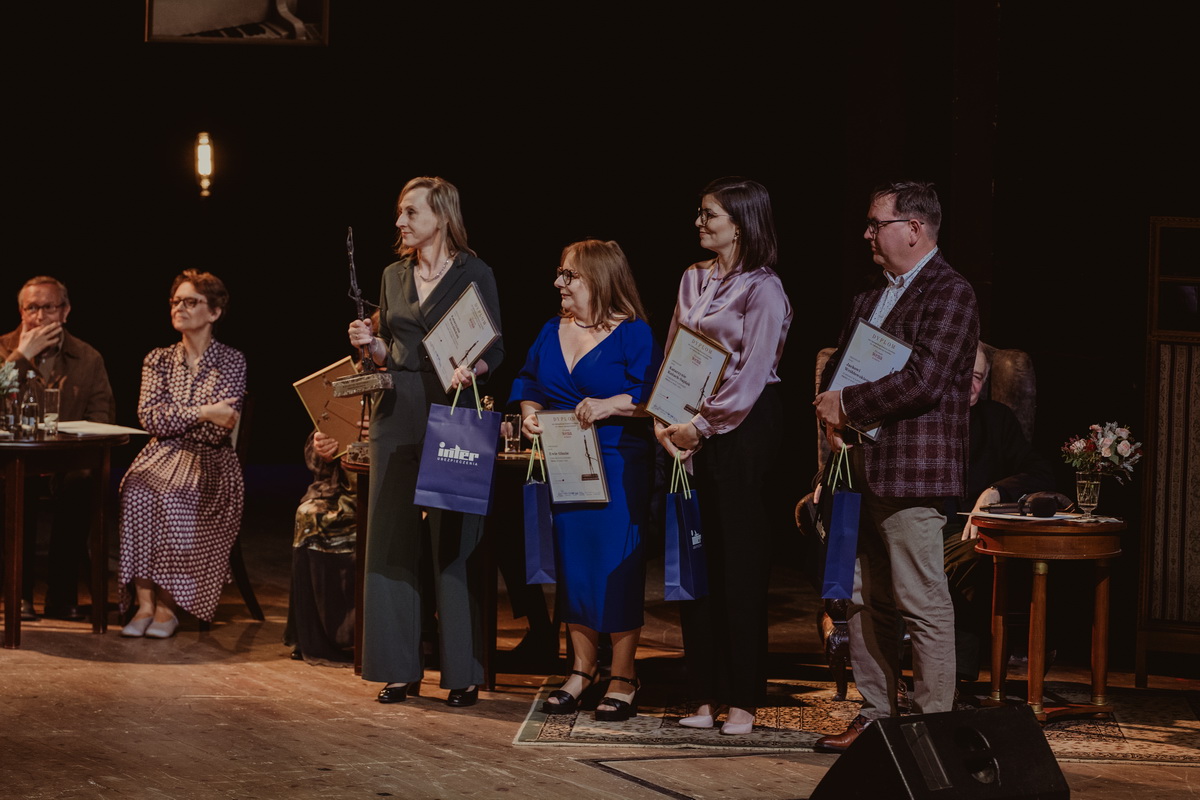 Na teatralnej scenie stoi grupa uśmiechniętych osób trzymają w w dłoniach upominki 