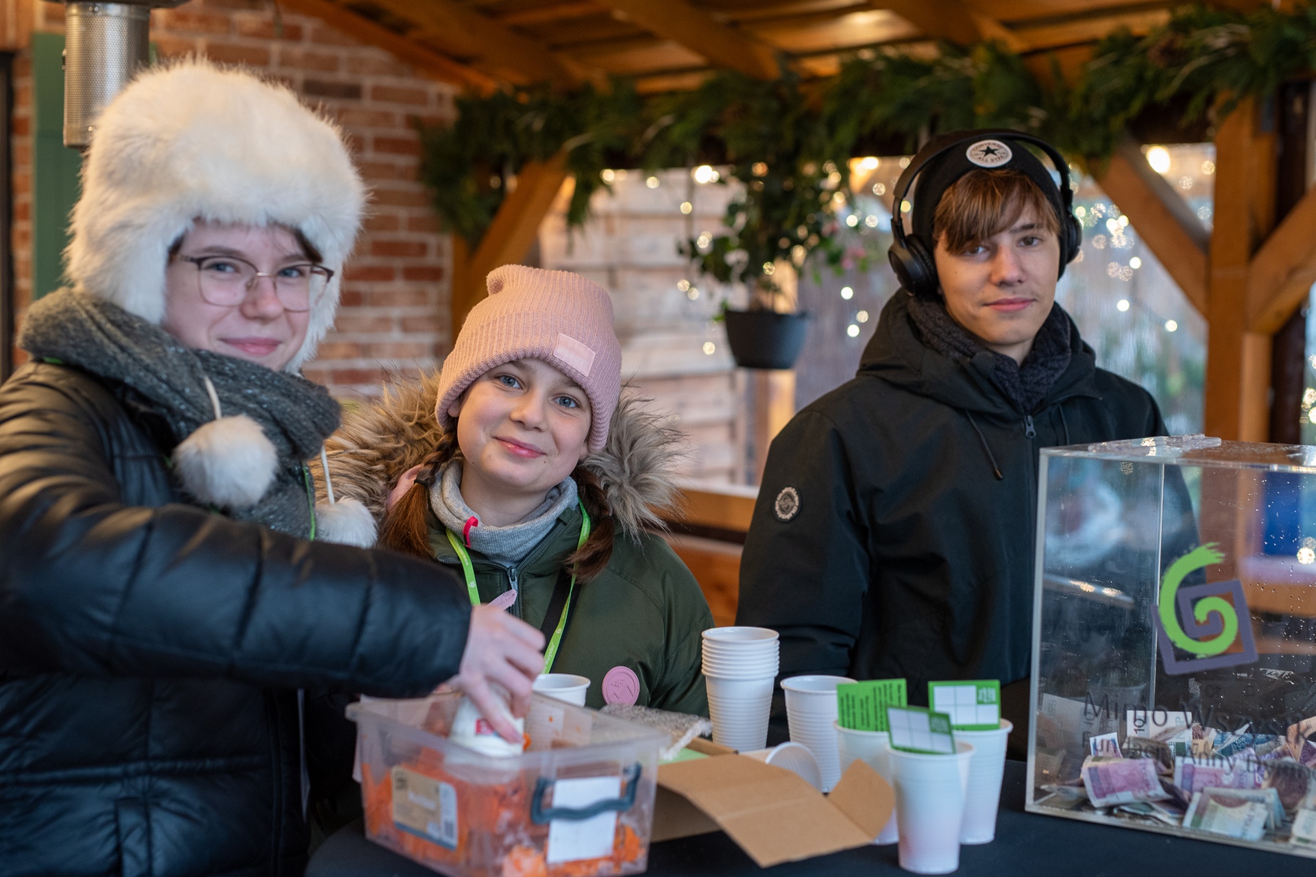 Trzy młode osoby pozują do zdjęcia. Widoczne są od popiersia w górę. Dwie dziewczyny po lewej stronie i chłopak po prawej. Wszyscy ubrani są w zimowe kurtki i czapki. Stoją pod dachem drewnianej altany. Przed nimi znajduje się stół. Na nim stoi przezroczysta puszka na darowizny pieniężne. Obok plastikowe kubeczki i pojemnik z warzywami. W tle widać ozdoby świąteczne i lampki.