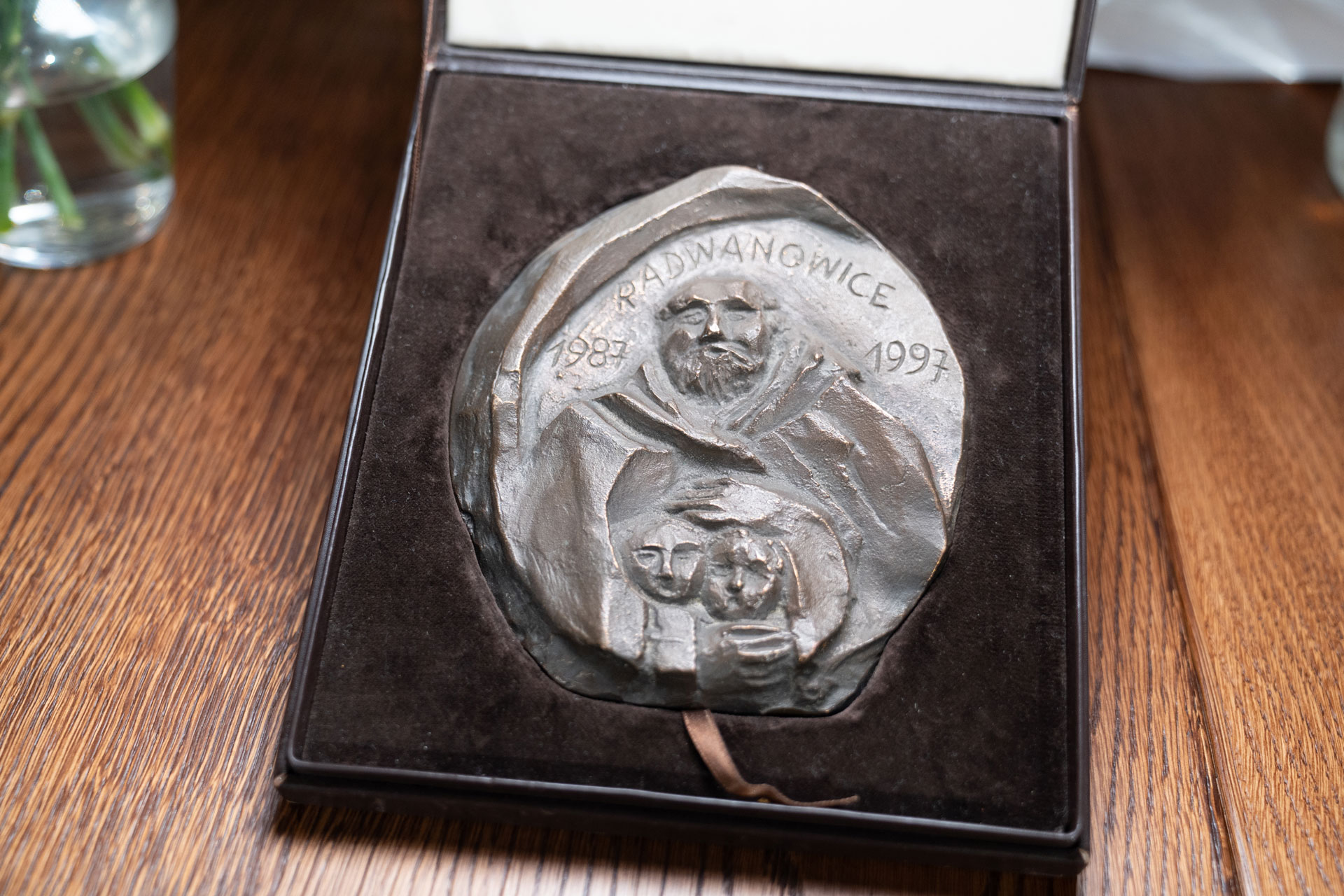 Na drewnianym stole leży ciemnobrązowy pokrowiec. Jest otwarty. W nim znajduje się duży medal. Wygląda jak wyrzeźbiony w metalu. Rzeźbienie przedstawia mężczyznę z brodą, w habicie, przytulającego dwoje dzieci. Na górze medalu wyrzeźbiono napis – od lewej: „1987 Radwanowice 1997”. W lewym górnym rogu zdjęcia widać na stole fragment przezroczystego wazonu z kwiatami.