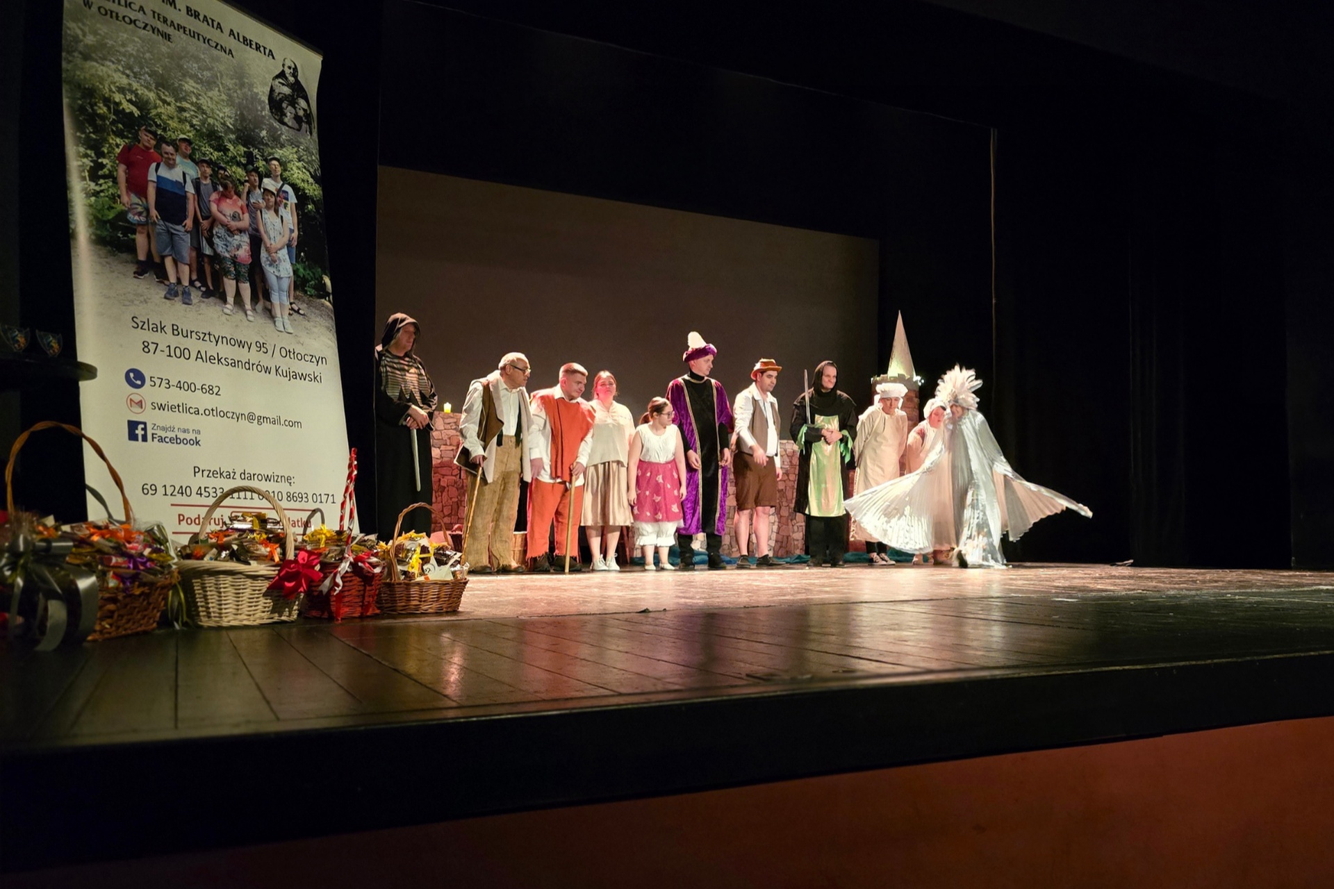 Grupa ludzi stoi na scenie teatralnej, w jej głębi. Są ubrani w kolorowe stroje. Stoją w linii, przodem do widowni. Po lewej stronie zdjęcia, na skraju sceny, stoją koszyki wiklinowe z kwiatami i wstążkami. Za nimi stoi wysoki roll-up z napisem: „Fundacja im. Brata Alberta. Świetlica Terapeutyczna w Otłoczynie”, ze zdjęciem grupy ludzi i danymi kontaktowymi.