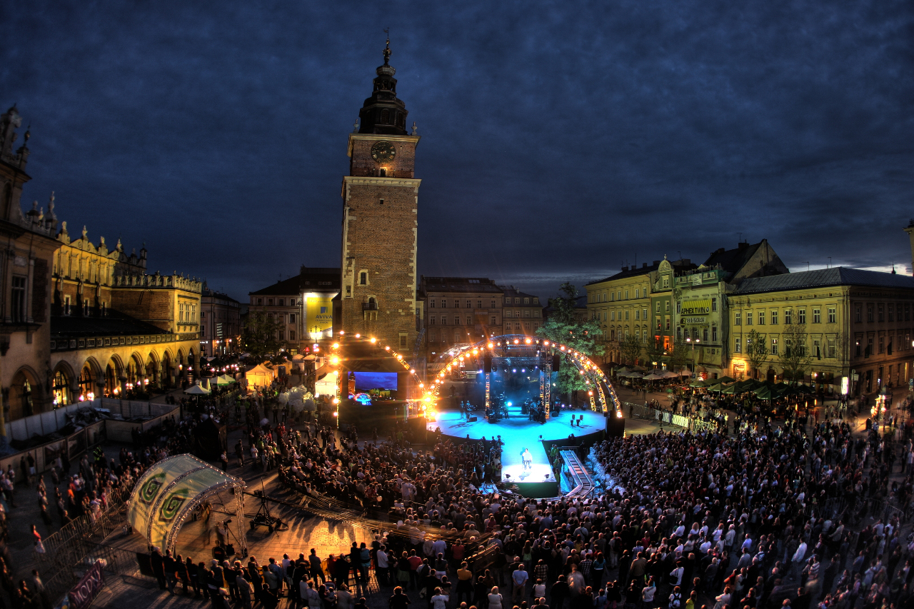 Pogrążony w nocy Rynek główny w Krakowie. Po lewej stronie Sukiennice, po prawej zabytkowe kamieniczki. Na wprost wieża Ratusza. Pod nią scena rozświetlona jasnymi reflektorami. 